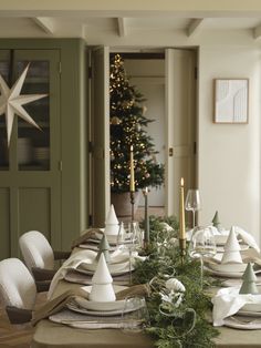 the table is set for christmas dinner with white dishes and silverware, greenery