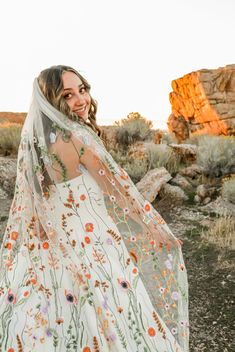 a woman standing in the desert wearing a white dress with flowers on it and a veil over her head