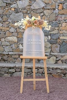 a sign with flowers on it sitting in front of a stone wall