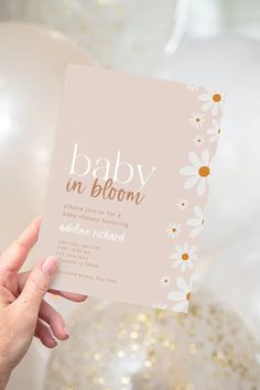 a person holding up a baby in bloom card next to white and gold balloons with daisies on them