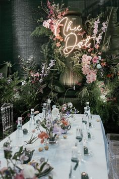 a long table with flowers and bottles on it in front of a neon sign that says live eat