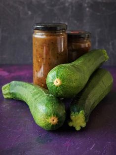 three cucumbers and two jars of mustard sit on a purple surface