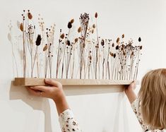 a woman holding up a piece of art with dried flowers on the wall behind her