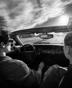 two people in a car driving down the road with sun shining through the clouds behind them