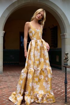 a woman in a yellow and white floral print dress standing on a brick walkway near an archway