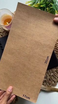 a person holding up a piece of brown paper on top of a white table next to a cup of tea