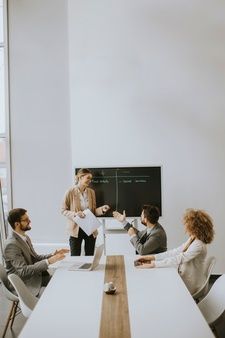 four people sitting at a table with papers in front of them and one person standing up