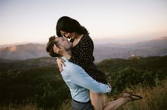 a man and woman hug each other while standing on top of a hill with mountains in the background