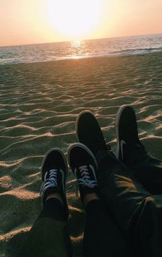 two people are sitting on the beach with their feet up in the sand as the sun sets