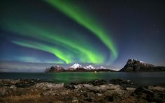 an aurora bore is seen over the mountains and water in this photo, with rocks on the foreground