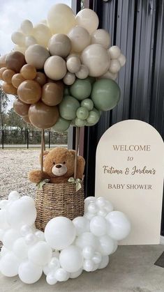 a teddy bear sitting on top of a basket filled with white and green balloons next to a welcome sign