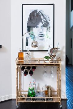 a gold bar cart with bottles and glasses on it in front of a framed photograph