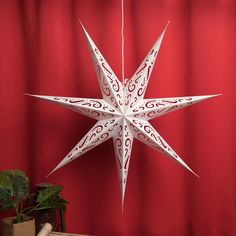 a white paper star hanging from a red curtain next to a plant and box on a table