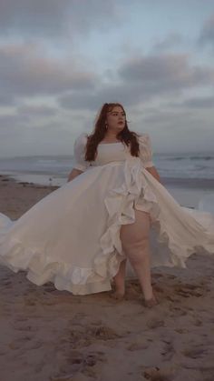 a woman in a white dress standing on the beach with her arms around her body