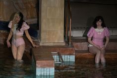 two women in pink swimsuits sitting on steps next to a pool with water