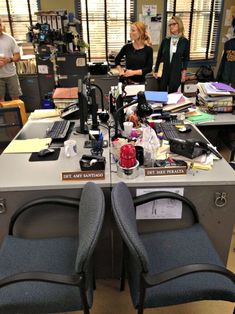 two office desks with chairs in front of them and people standing around the room