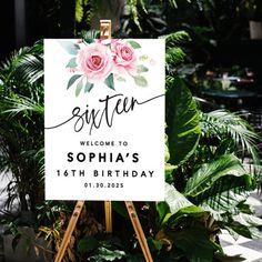 a welcome sign with pink flowers on it in front of some plants and greenery