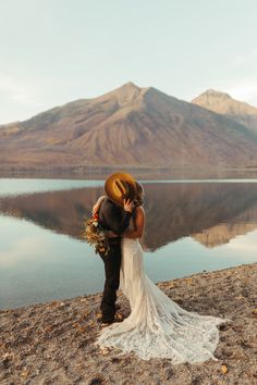 a bride and groom kissing on the shore of a lake with mountains in the background