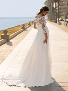 a woman in a white wedding dress is standing on the sidewalk near the water and looking down