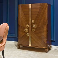 a wooden cabinet sitting next to a pink chair in front of a blue wall with gold trim