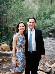 a man and woman standing next to each other in front of a river with rocks