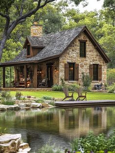 a stone house sitting next to a lake surrounded by trees and grass with two chairs on the lawn