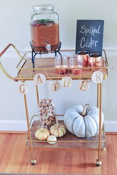 a gold serving cart with candles and pumpkins on it, along with a chalkboard sign that says spiced cider