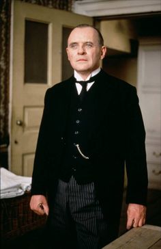 a man in a suit and tie standing next to a wooden table with his hands on his hips