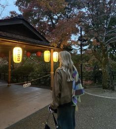 a woman is walking with her bike in front of a pavilion at dusk, and lanterns are hanging from the roof