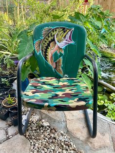 a chair with a fish painted on it sitting next to some plants and rocks in a garden