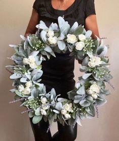 a woman is holding a wreath with white flowers and greenery in front of her