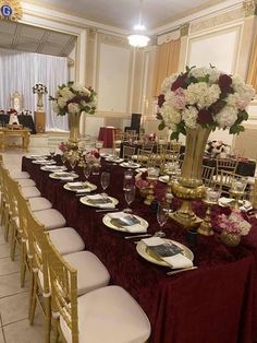 a long table is set up with white and red flowers in gold vases, plates and silverware