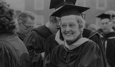 black and white photograph of woman in graduation cap and gown smiling at camera with other people behind her