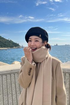 a woman wearing a hat and scarf standing next to the ocean with her hand on her mouth