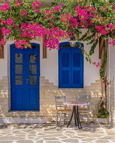 a table and chairs under blue shuttered windows with pink flowers on the outside wall