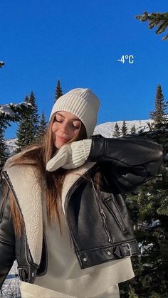 a woman wearing a black leather jacket and white sweater is standing in front of some trees