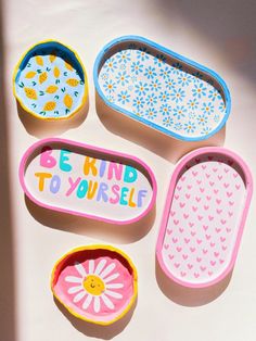 four small trays with painted designs on them sitting on a white surface, one has the words be kind to yourself