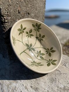 a white bowl sitting on top of a rock next to the ocean with green leaves painted on it