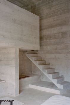 a bedroom with concrete walls and stairs leading up to the second floor, next to a bed