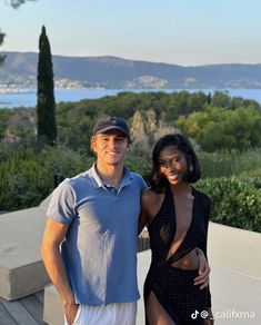 a man and woman standing next to each other in front of trees with water behind them