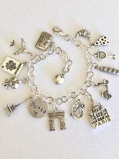 "Adorable Paris charm bracelet featuring 13 detailed silver charms of travel to Paris, classic landmarks, artist quarter, food and drink. Hand stamped 1/2\" 18 aluminum disc says \"Paris ❤️\". Memories of a visit abroad living \"la bonne vie\" in the magical city of Paris! Charms include: suitcase, plane, passport, fleur de lis, Eiffel Tower, Arc de Triumph, artist palette, Sacre Coeur church, Vespa scooter, baguette, cheese, wine, and croissant. Charms are antique silver, pewter and silver tone Silver Engraved Jewelry For Travel, Nickel-free Silver Charms Souvenir, Nickel-free Silver Charms For Souvenir, Silver Charms Jewelry Souvenir, Silver Metal Jewelry For Travel, Adjustable Silver Jewelry For Travel, Silver Charm Bracelet As Souvenir, Silver Symbolic Jewelry Souvenir, Silver Nickel-free Bracelet Souvenir