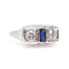 This lovely ring, an original Art Deco piece from the 1920s, has two bright set European Cut diamonds separated by a vertically oriented French Cut sapphire baguette. The shank is beautifully engraved and the undercarriage has swirl cutouts. The top of the ring measures 7mm wide, about the width of a large wedding band, and this would make an ideal engagement ring, with the two diamonds representing "You" and "I."DETAILS________________METAL: 14K White GoldDIAMONDS: 2x 4mm European Cut Diamond ( Art Deco Three Stone Platinum Jewelry, Art Deco Three Stone Ring For Formal Occasions, Vintage Sapphire Three Stone Ring For Formal Events, Collectible Art Deco Platinum Sapphire Ring, Vintage Three Stone Sapphire Ring For Formal Events, Vintage Three Stone Sapphire Ring For Formal Occasions, Collectible Art Deco Sapphire Ring, Art Deco Platinum Sapphire Ring With 17 Jewels, Art Deco Platinum Sapphire Ring