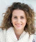 a woman with curly hair wearing a white scarf and plaid shirt smiling at the camera