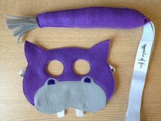 a purple cat mask with grey eyes and tail on a table next to a white lanyard