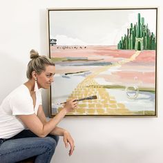 a woman sitting on a stool in front of a painting holding a paintbrush and looking at it