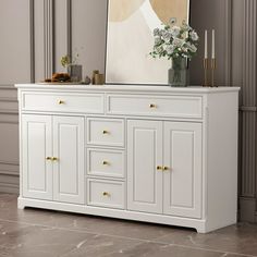 a white dresser with gold handles and drawers in front of a painting on the wall