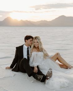 a man and woman are sitting on the ground in white dress clothes, with mountains in the background