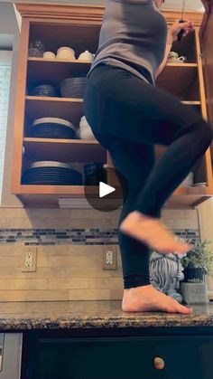 a woman standing on top of a kitchen counter next to an open cupboard filled with dishes