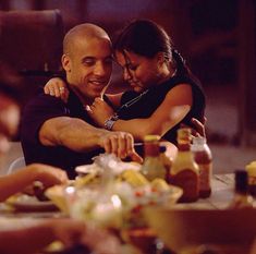 a man and woman hugging each other while sitting at a table with food on it