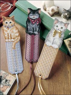 three cross stitch bookmarks with cats on them sitting on a table next to books